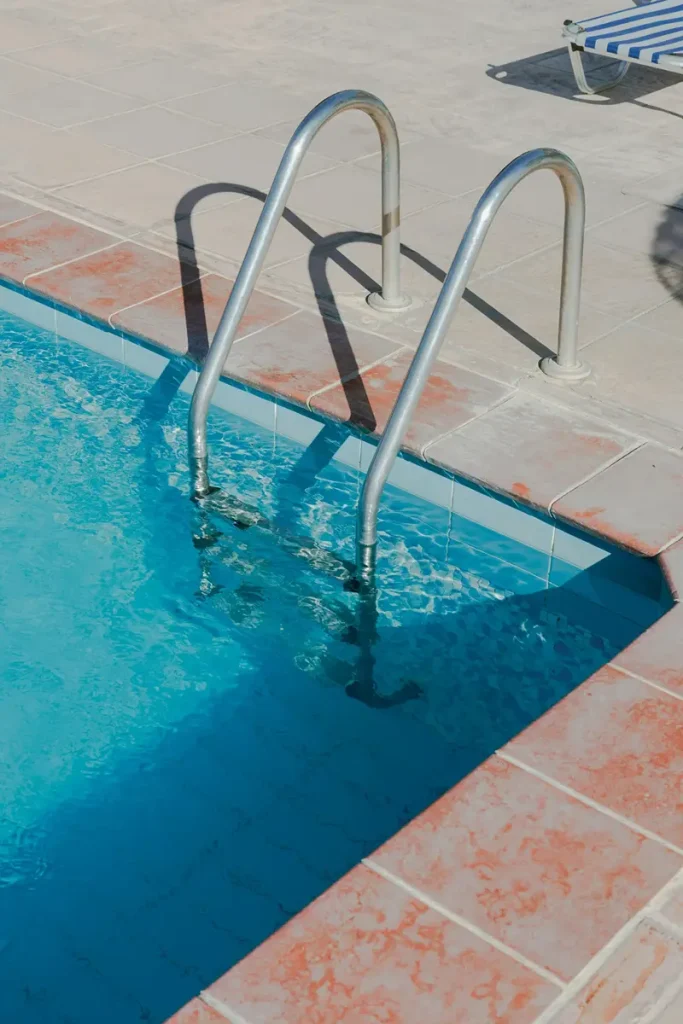 Close-up of dirty pool coping tiles and a metal ladder showing slick organic buildup and water stains before a pressure washing treatment.