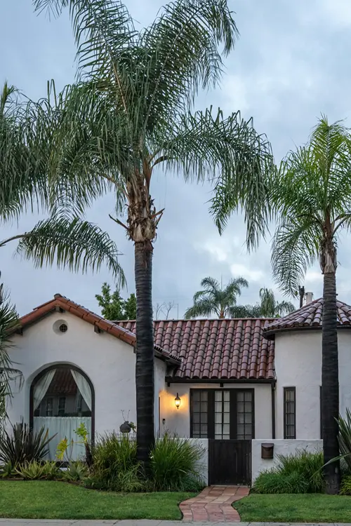 A luxury white stucco residential estate with immaculate dark clay roofing tiles and prominent green palm trees in the front yard.